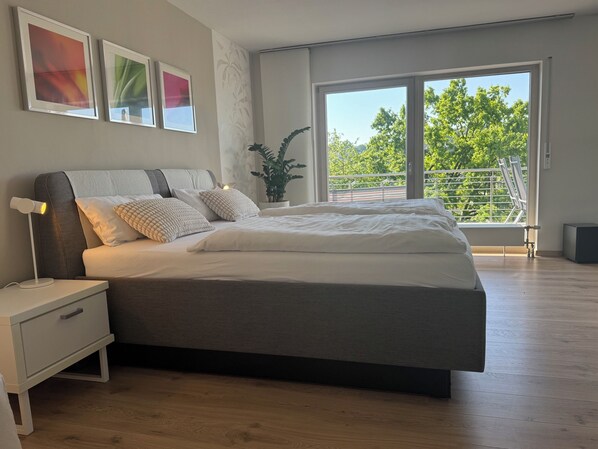 A modern bedroom with a large bed, bedside tables, a nightstand, a potted plant, and a view of trees through a large window. - Modern, well-equipped 5* vacation apartment on the sunny side of Linz am Rhein (Linz am Rhein)
