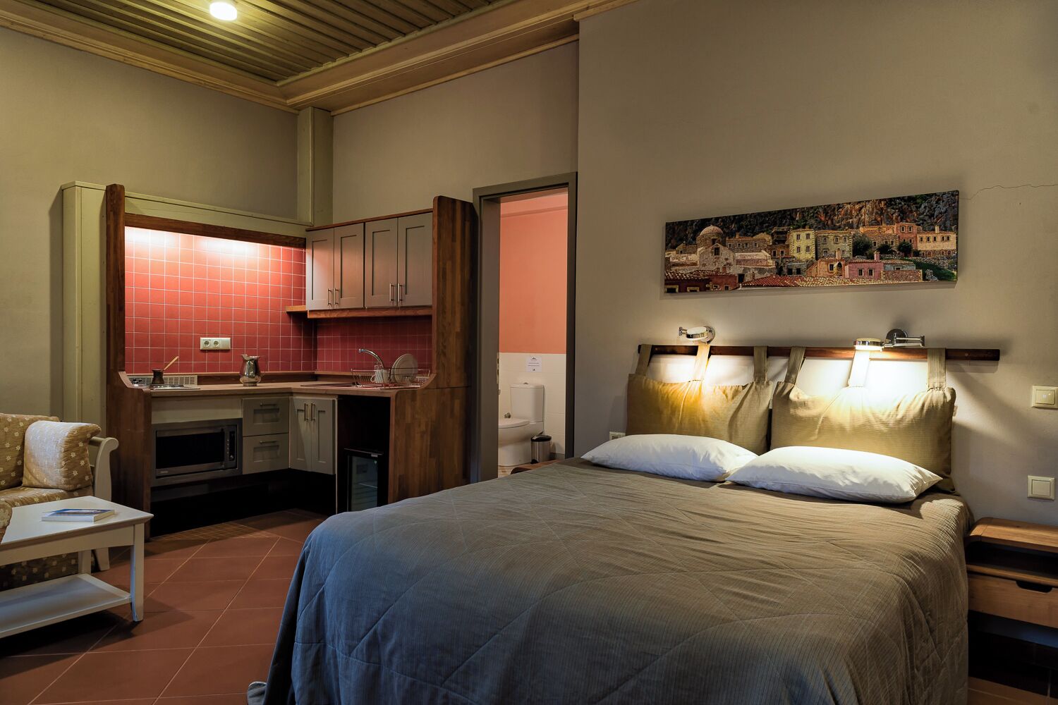 A compact hotel room with a bed, bedside tables, a small kitchenette, and a painting on the wall.