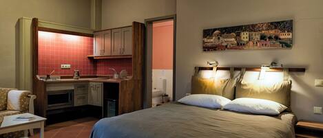 A compact hotel room with a bed, bedside tables, a small kitchenette, and a painting on the wall.