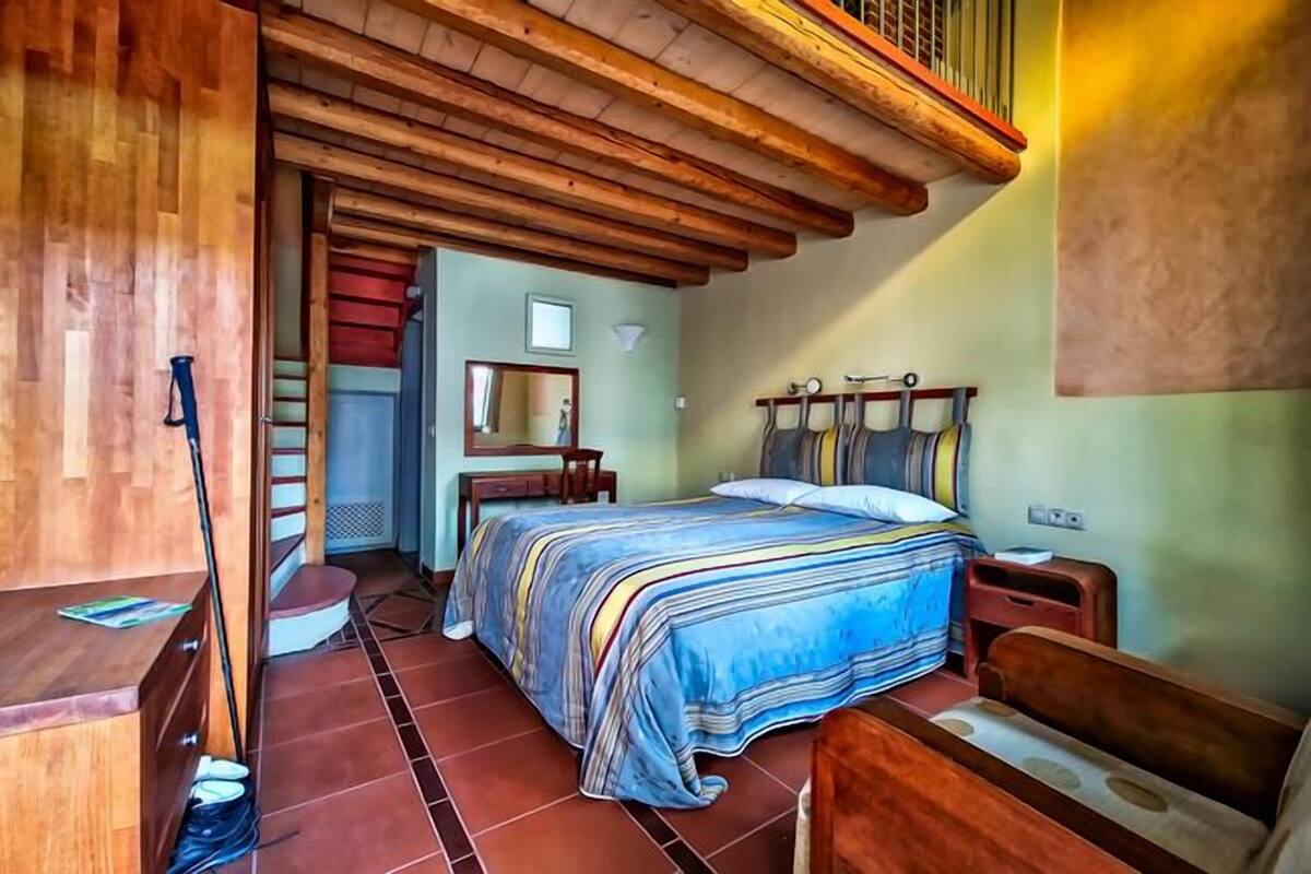 Une chambre avec un plafond en bois, un lit avec une literie à rayures, une table de chevet et un banc en bois.