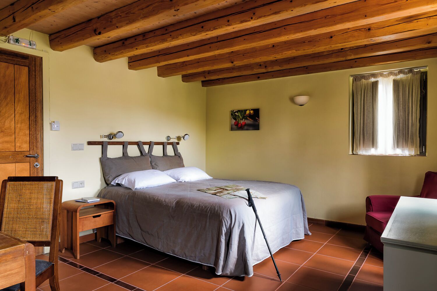 A bedroom with a wooden ceiling, a bed with a headboard, a nightstand, a chair, a table, and a window with curtains.