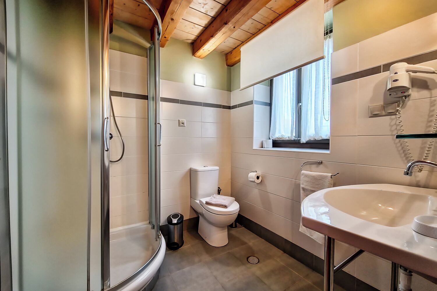 Un baño con ducha envuelta en cristal, inodoro, lavabo y ventana con cortinas.