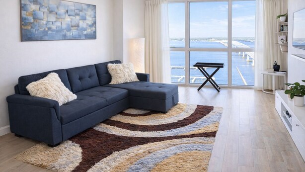 A modern living room with a dark blue sectional sofa, a patterned rug, a large window with a view of the sea, and a small table with a lamp. - Have a Relaxing getaway from day by day stress
Cozy apartment with stunning view (Fort Myers)