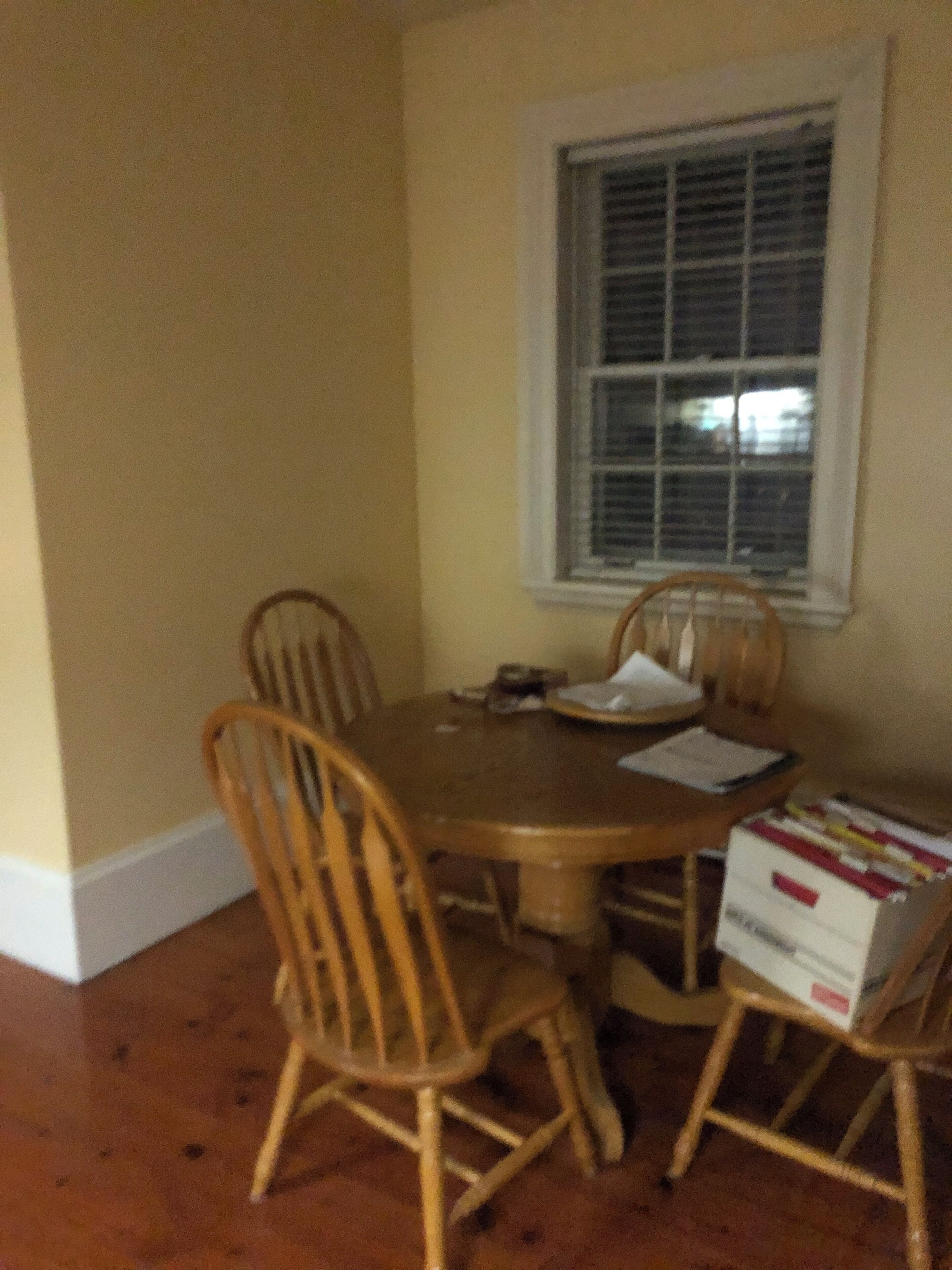 Un coin repas avec une table ronde et quatre chaises, une fenêtre avec des stores et une pile de livres posée sur le sol.