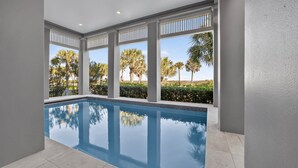A swimming pool with clear blue water, surrounded by white columns and large windows offering views of palm trees and a clear sky. - New Beachfront Listing - Heated Pool, Golf Cart, Elevator, Steps to Best Beach (Galveston)