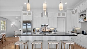 A modern kitchen with white cabinetry, a large island with bar stools, pendant lighting, and a stainless steel refrigerator. - New Beachfront Listing - Heated Pool, Golf Cart, Elevator, Steps to Best Beach (Galveston)