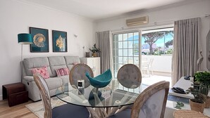 Uma sala de estar moderna com uma mesa de jantar de vidro, sofá cinza e vista para o exterior através de grandes janelas.