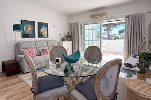 Een moderne woonkamer met een glazen eettafel, een grijze bank en een uitzicht op de omgeving door grote ramen.