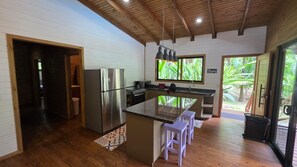 A modern kitchen with a stainless steel refrigerator, a built-in island with bar stools, a window with a view of greenery, and a wooden ceiling.