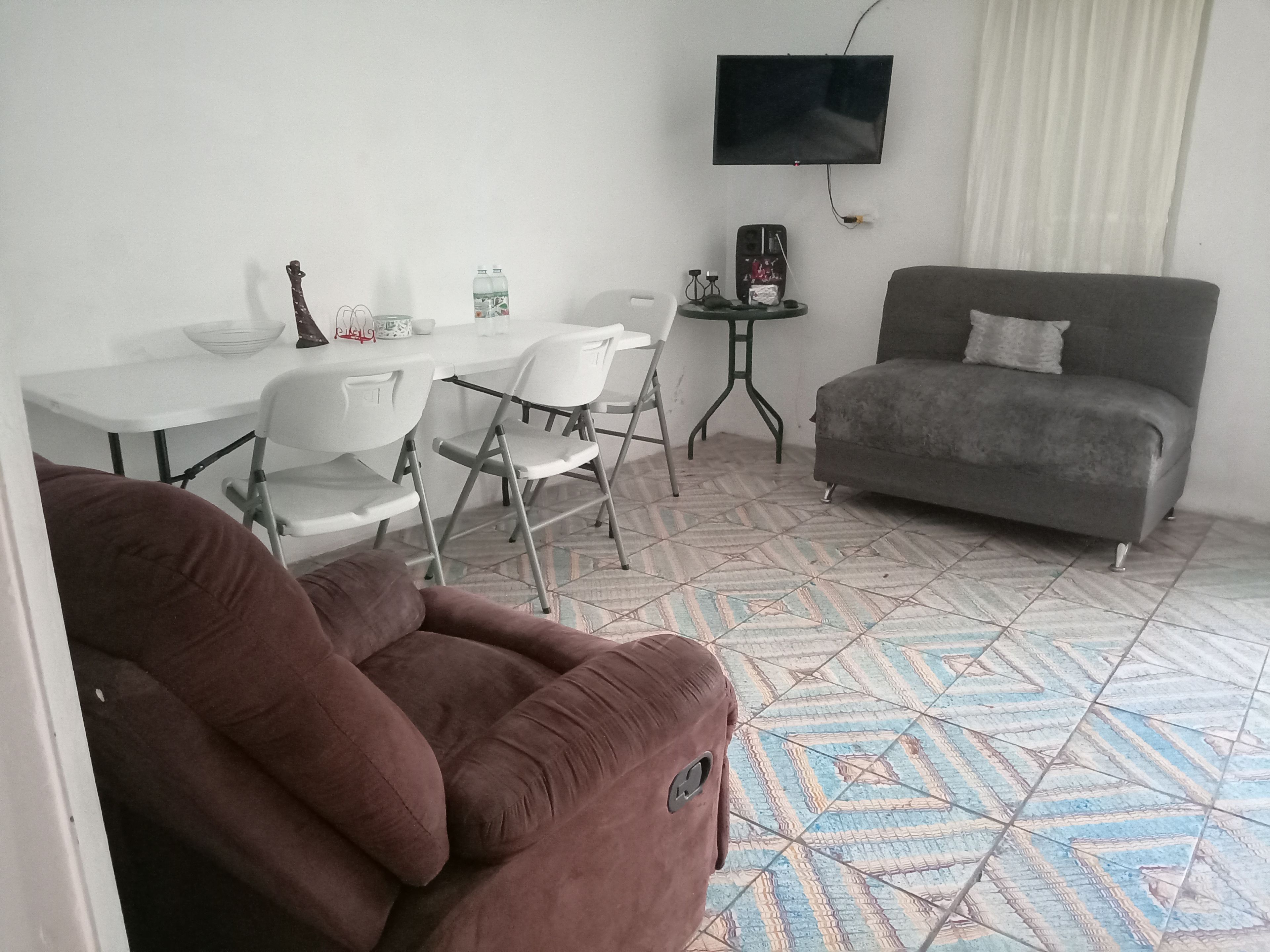 Zona d'estar amb sofà, taula de dinar, cadires, televisor fixat a la paret i terres de rajoles amb dibuixos.