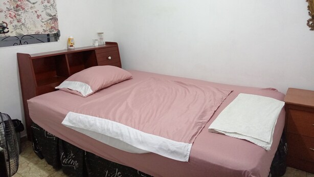 Un letto singolo con copriletto rosa e cuscino bianco, un comodino in legno con una lampada, un tavolino con una bottiglia e un bicchiere, e una parete con una tenda a fiori.