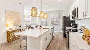 A modern kitchen with white cabinetry, a large island, pendant lighting, and a dining area with a table and chairs. - Sea La Vie Corner Condo at Gulf Place Wraparound Balcony & Beach Access (Santa Rosa Beach)