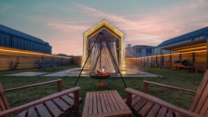 Een terras met houten stoelen en een tafel, een vuurkorf en een glazen structuur met dak.