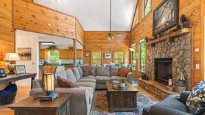 A cozy cabin interior with a stone fireplace, wooden walls, and a large sofa in the living area. - Breathtaking Views for Family & Friends to Gather (Clarkesville)