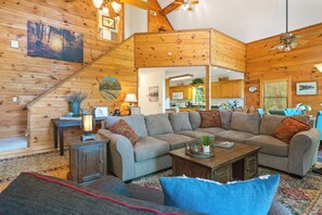 A spacious living room with a large sectional sofa, a wooden staircase, and a kitchen visible through an open doorway. - Breathtaking Views for Family & Friends to Gather (Clarkesville)