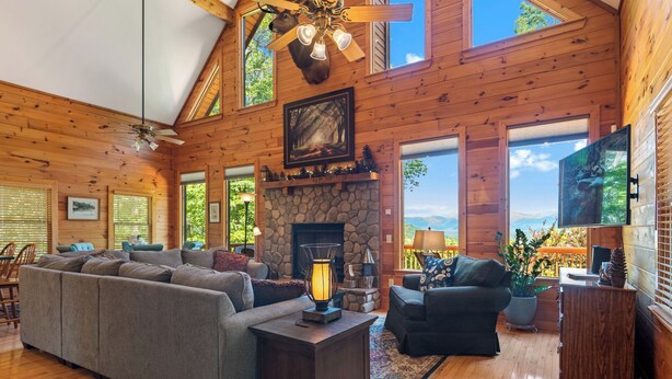 A spacious living room with a stone fireplace, wooden ceiling, and large windows offering a view of the outdoors. - Breathtaking Views for Family & Friends to Gather (Clarkesville)