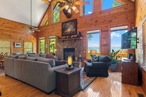 A spacious living room with a stone fireplace, wooden ceiling, and large windows offering a view of the outdoors. - Breathtaking Views for Family & Friends to Gather (Clarkesville)
