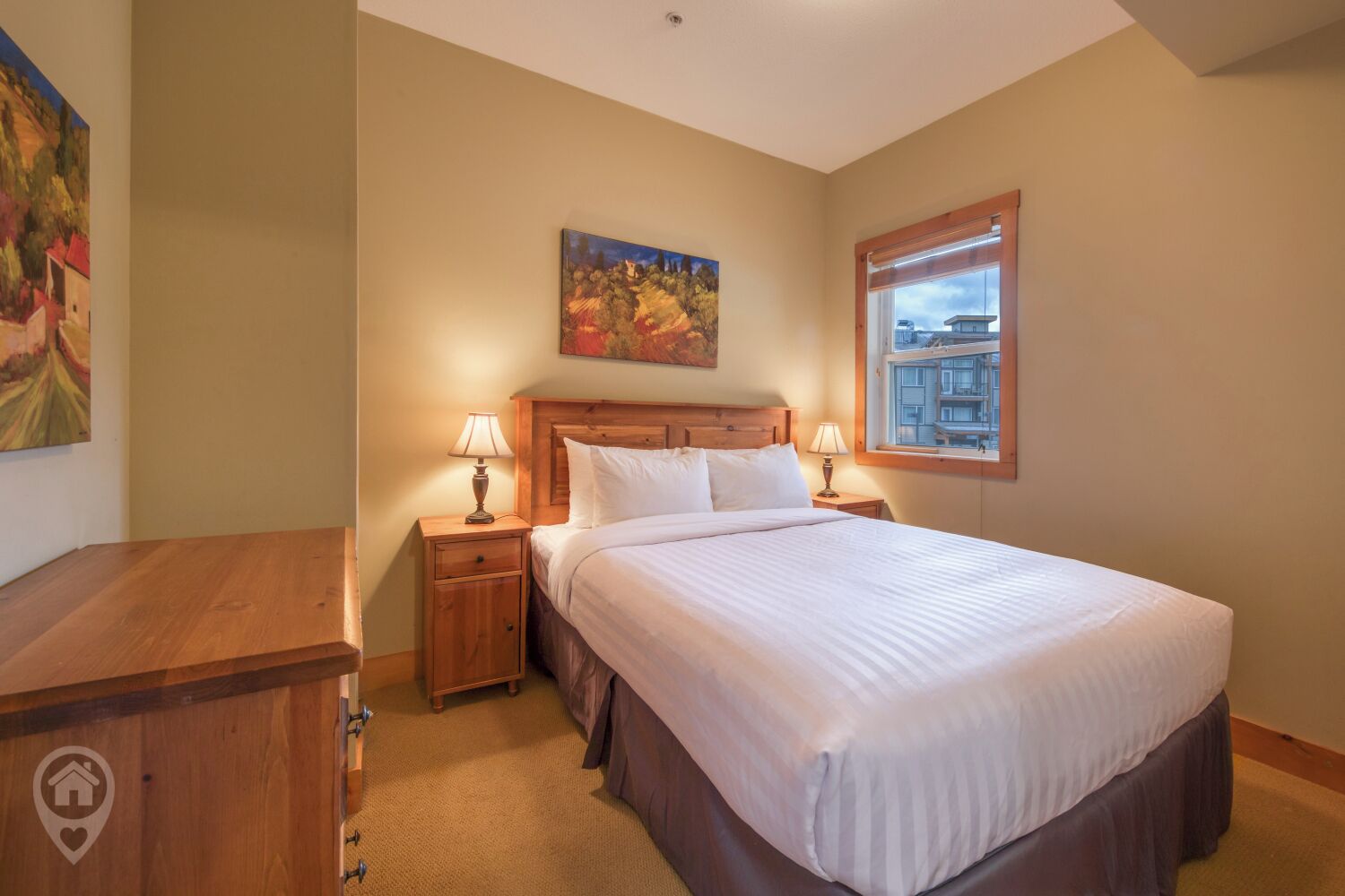 A bedroom with a large bed, wooden headboard, two bedside tables with lamps, a painting on the wall, and a window with a view of buildings.