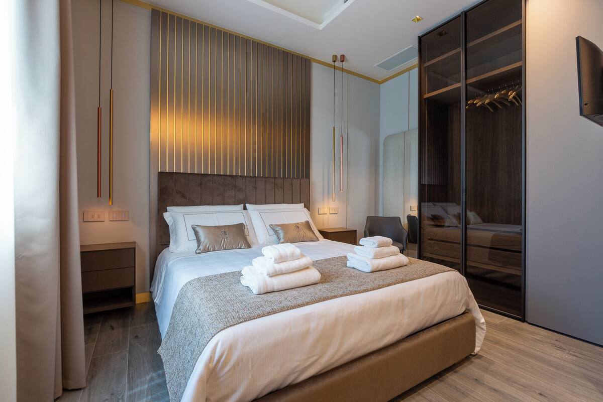 A modern hotel bedroom with a large bed, bedside tables, a wardrobe, and a mirror.