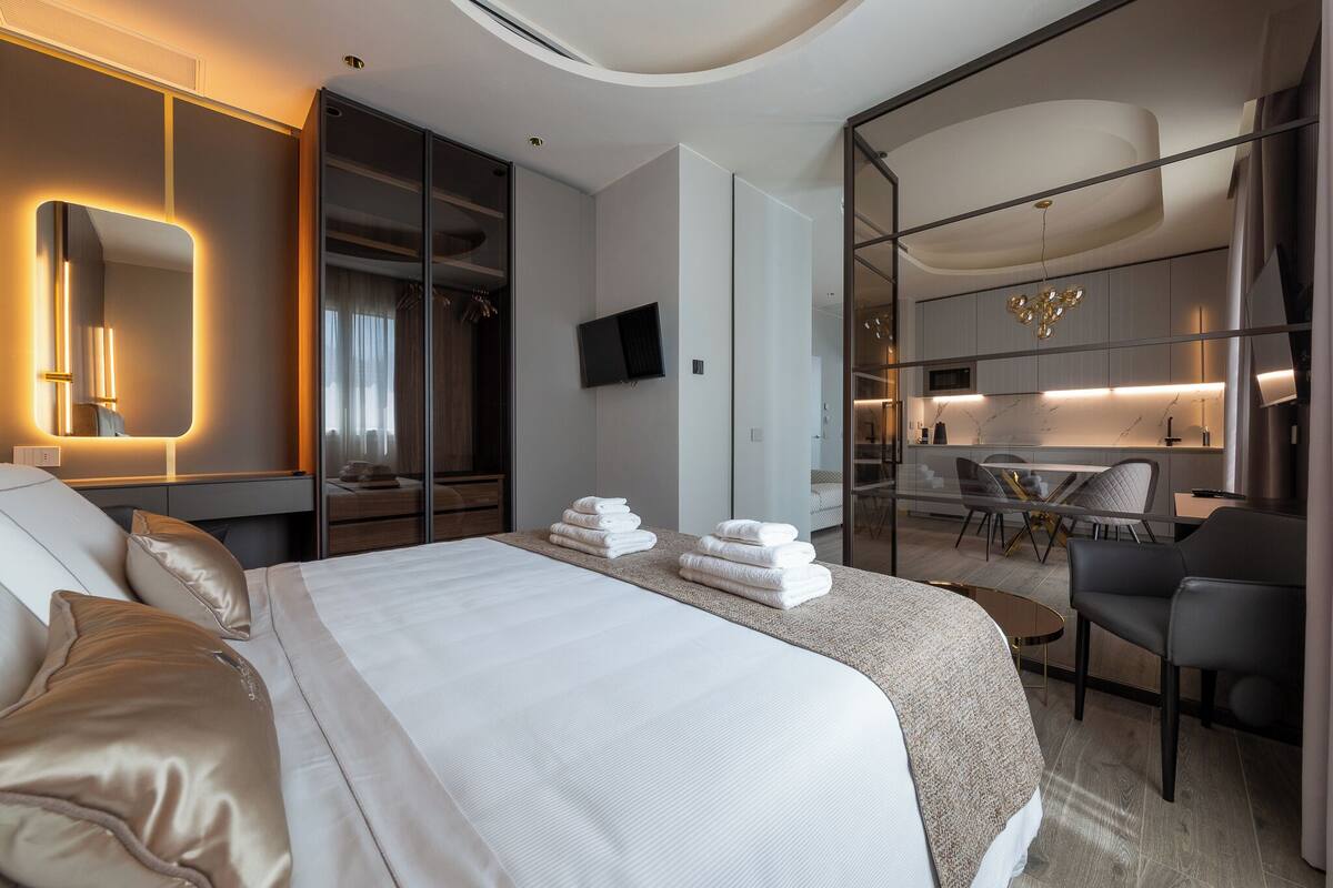 A modern hotel room with a large bed, a mirror, a TV, and a seating area with a table and chairs.