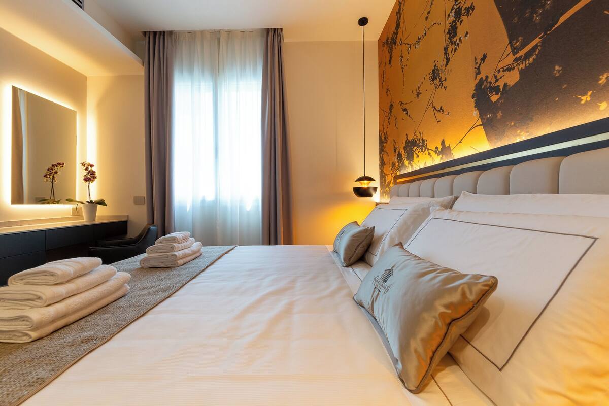 A hotel room with a large bed, a bedside table with a lamp, a mirror, and a view of the outdoors through the window.