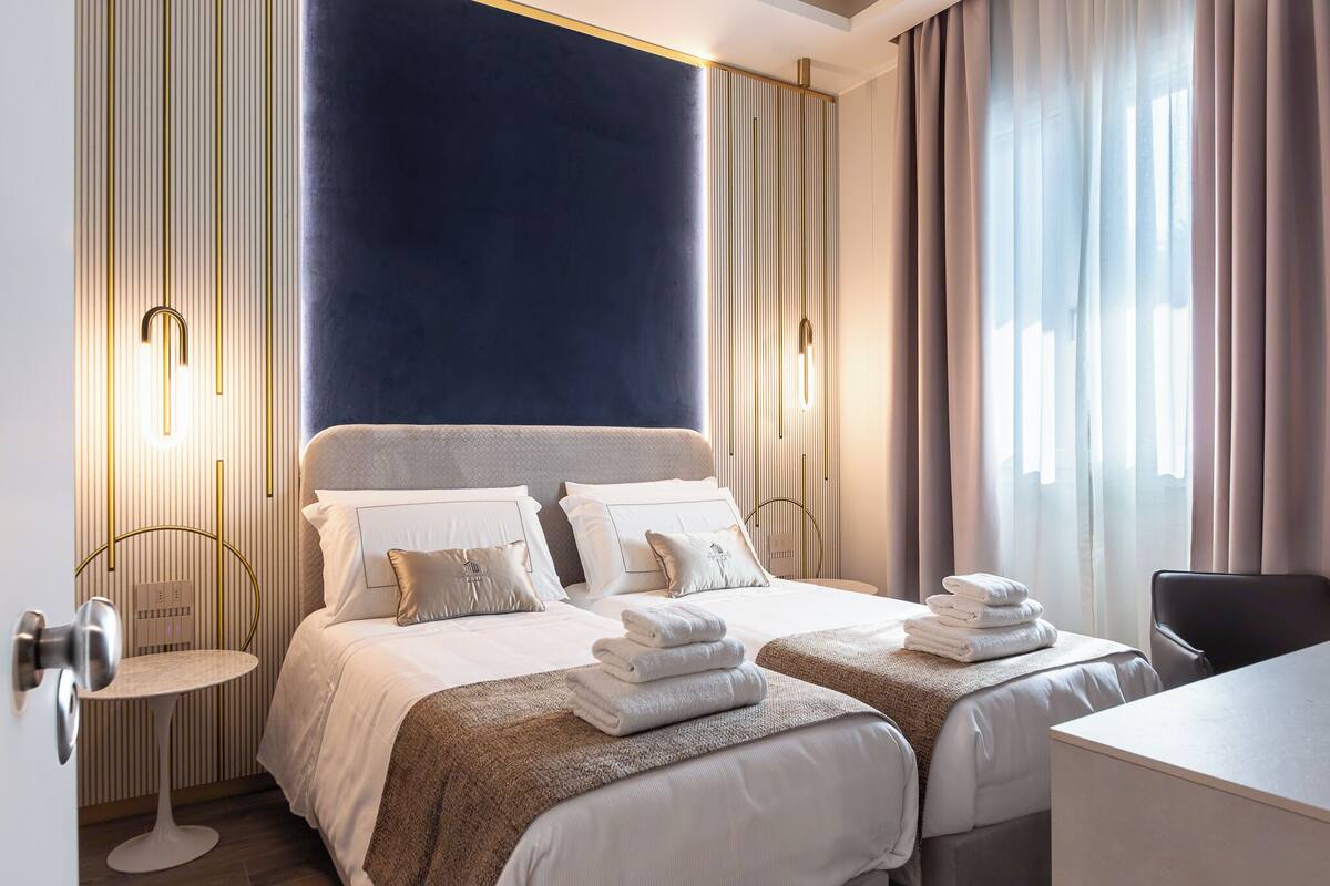 A hotel room with two beds, a desk, and a chair. There are towels on the beds and a table with a lamp.
