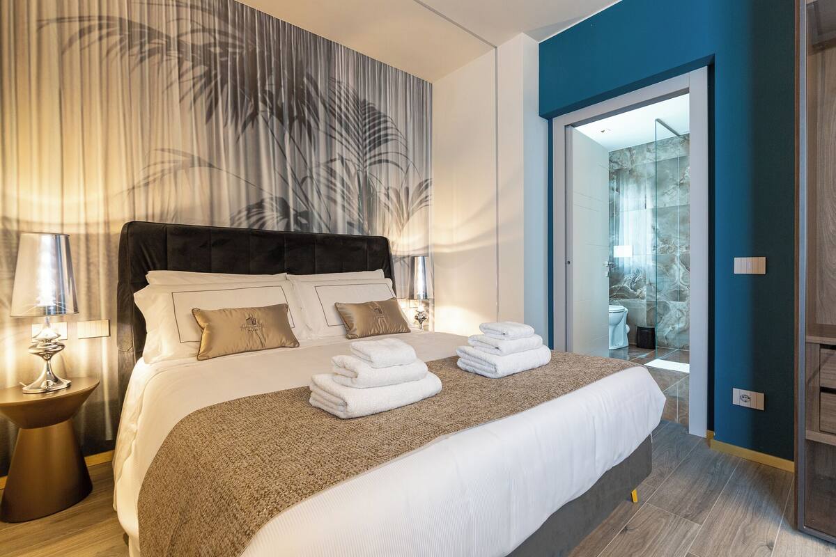 A hotel room with a large bed, a bedside table with a lamp, and a view of the bathroom through a glass door.