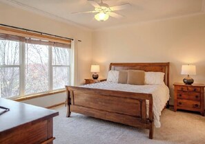 Un dormitorio con cama de madera, dos mesitas de noche con lámparas, un ventilador de techo y un ventanal con persianas.
