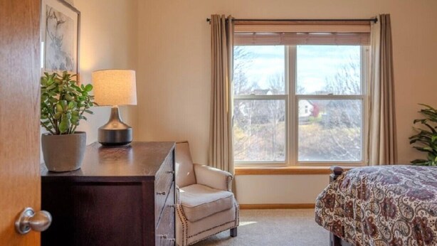 A bedroom with a bed, a chair, a desk with a lamp, a window with curtains, and a plant. - Spacious Modern 5-Bed Home –  Near Twin Cities (Rosemount)