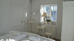 Een witte slaapkamer met een bed, een stoel, een bureau, een radiator en een raam met uitzicht op bomen.