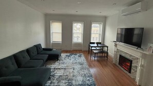 A living room with a sectional sofa, a fireplace, a TV, and three windows. - Cozy Harlem Brownstone 1BR Washer & Dryer (New York)
