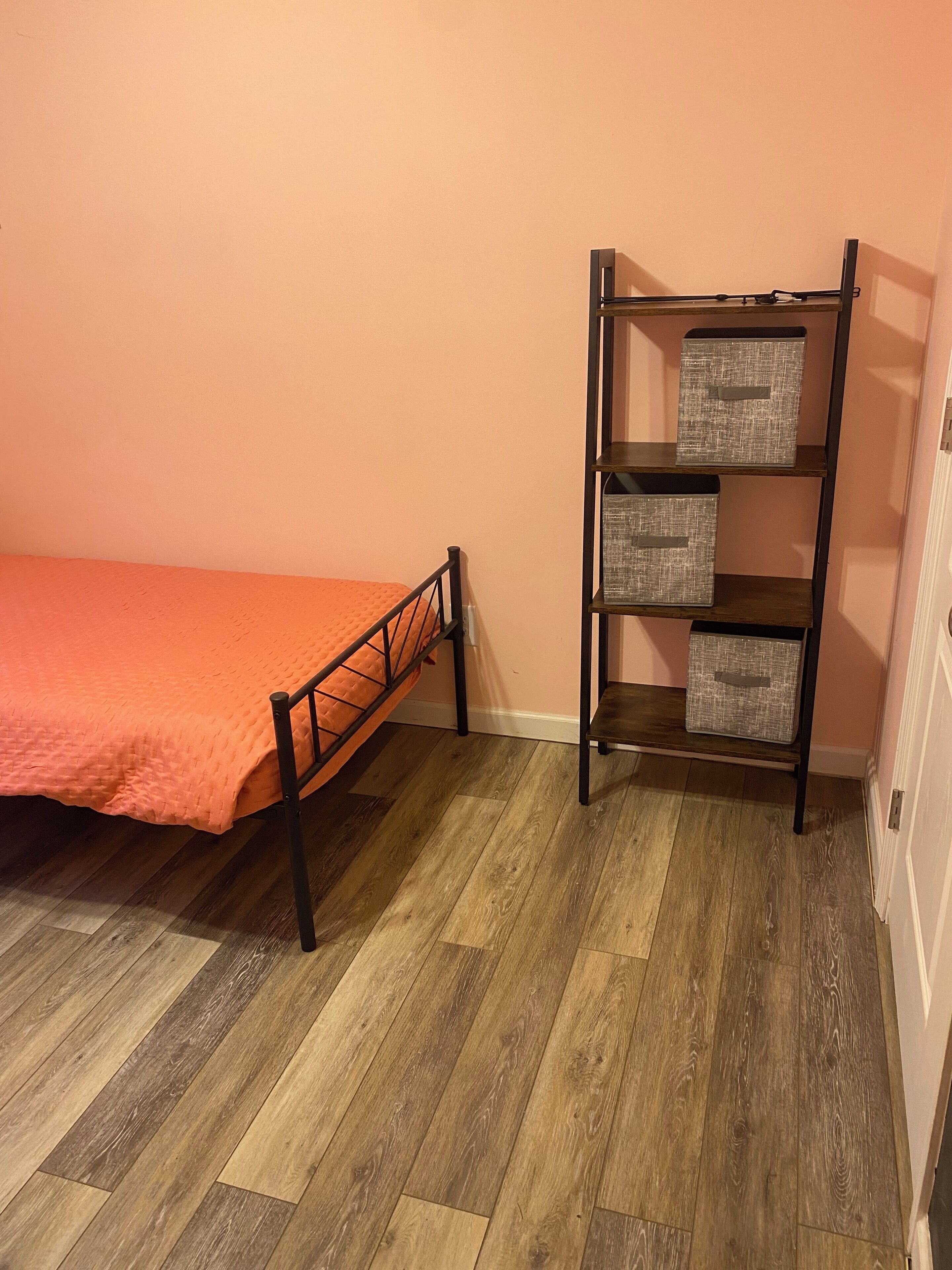 Uma cama de solteiro com colcha laranja, estrutura de metal e chão de madeira.