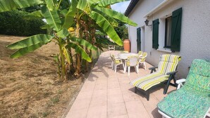 Un patio con una mesa y sillas, una silla lounge a rayas y una casa con contraventanas verdes.