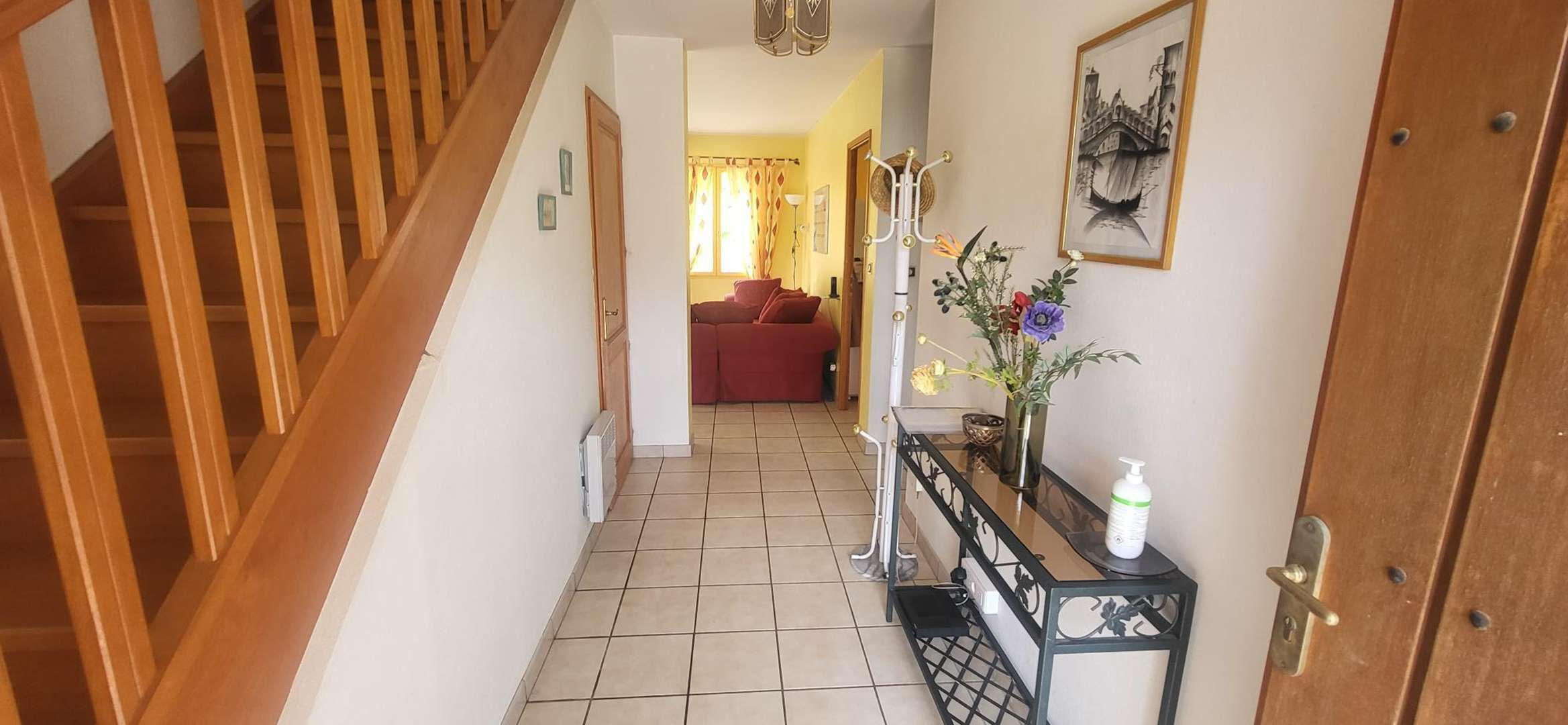 Een hal met een houten trap, een betegelde vloer, een rode bank op de achtergrond en een console tafel met een bloemenvaas en een fles.