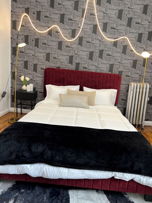 A bed with a red headboard, white bedding, and a black throw blanket. There is a bedside table with a vase of flowers and a lamp. The wall has a geometric pattern.
