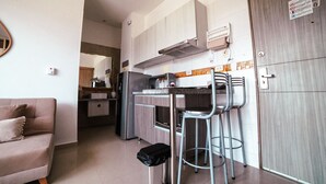 Et kompakt køkken med en central ø, indbygget ovn og køleskab. Der er spiseplads med to barstole og et lille bord. En sofa er synlig i forgrunden.