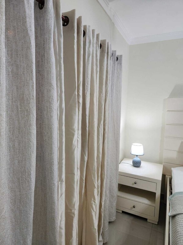 Een slaapkamer met een wit bed, een nachtkastje met een lamp en een raam met dunne gordijnen.
