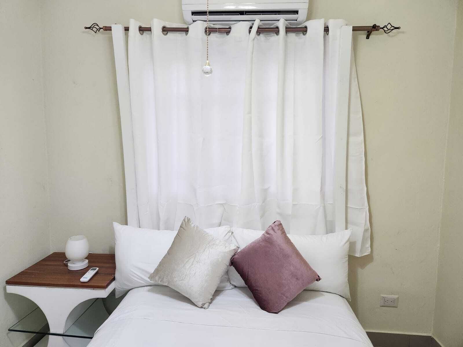 A hotel room with a bed, white curtains, a bedside table with a lamp, and a wall-mounted air conditioner.