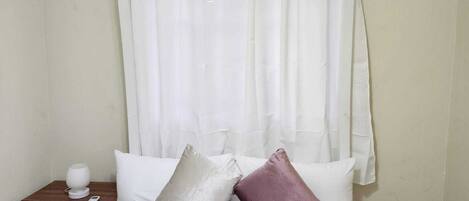 A hotel room with a bed, white curtains, a bedside table with a lamp, and a wall-mounted air conditioner.
