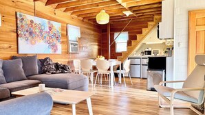 A cozy cabin interior with a wooden ceiling, a dining area, a kitchen, and a living room furnished with a sofa, chairs, and a coffee table.