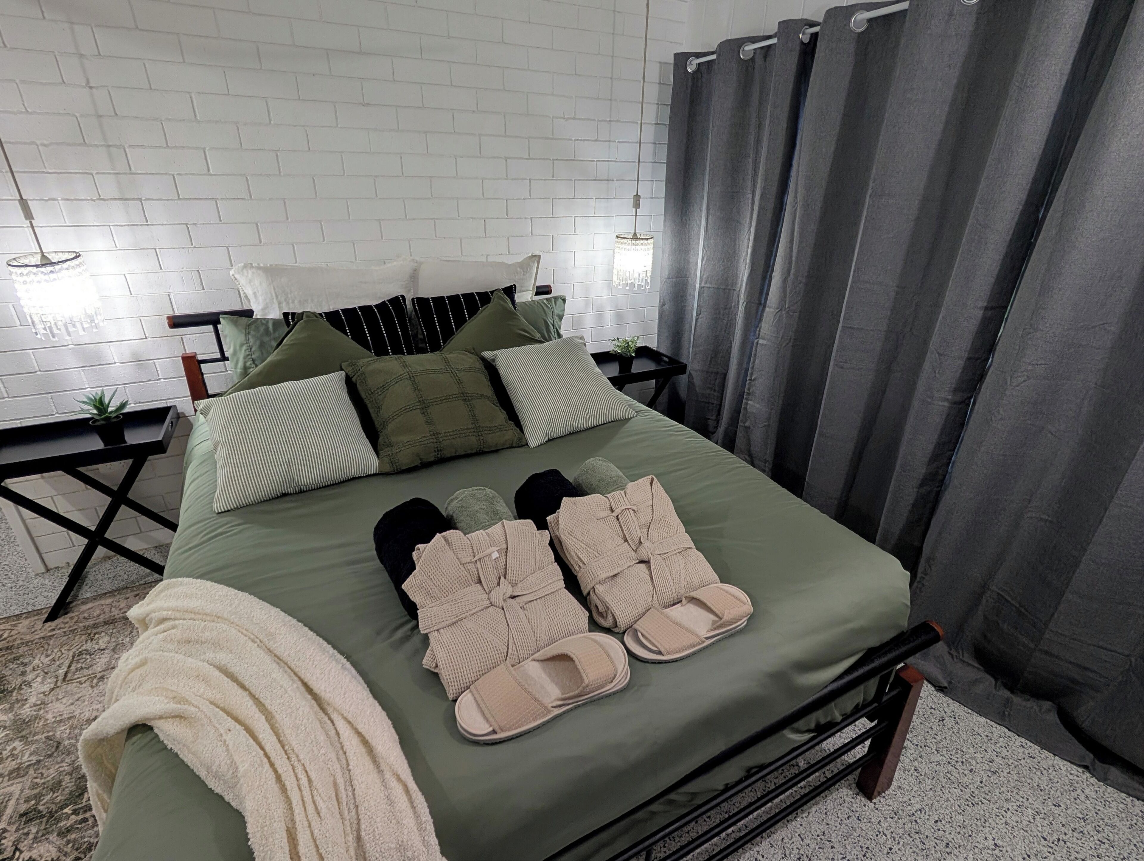 Ein ordentlich gemachtes Bett mit einer grünen Tagesdecke, mehreren Kissen und einem Paar Hausschuhen.
