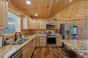 A kitchen with wooden cabinets, stainless steel appliances, a granite countertop, and a window with blinds. - Majestic Mountain Views • Hot Tub • Chalet Village (Gatlinburg)