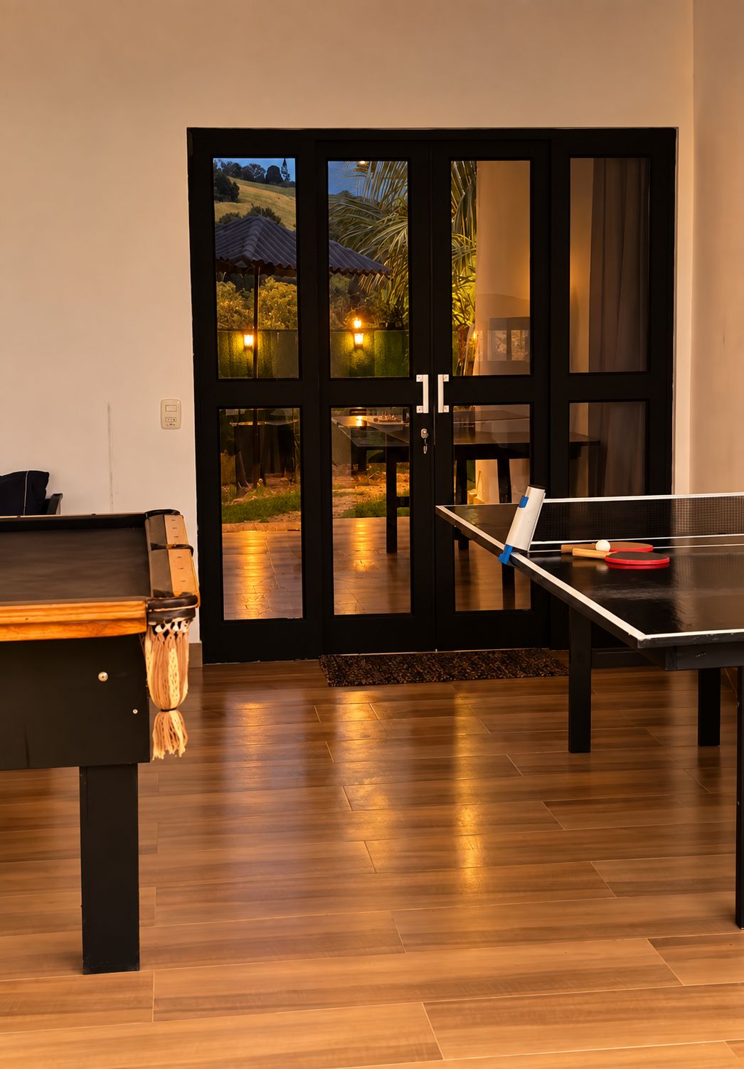 A room with a ping pong table, a pool table, and a view through a glass door.