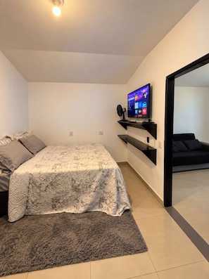 Ett modernt sovrum med en säng, en tv på ett stativ, en fläkt och en soffa.