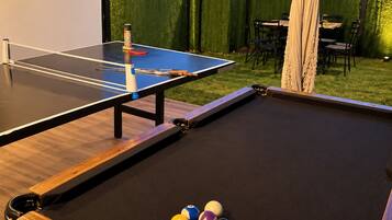 Ett biljardbord med bollar och ett bord för bordtennis i ett takförsett utomhusområde med utsikt över kullar.