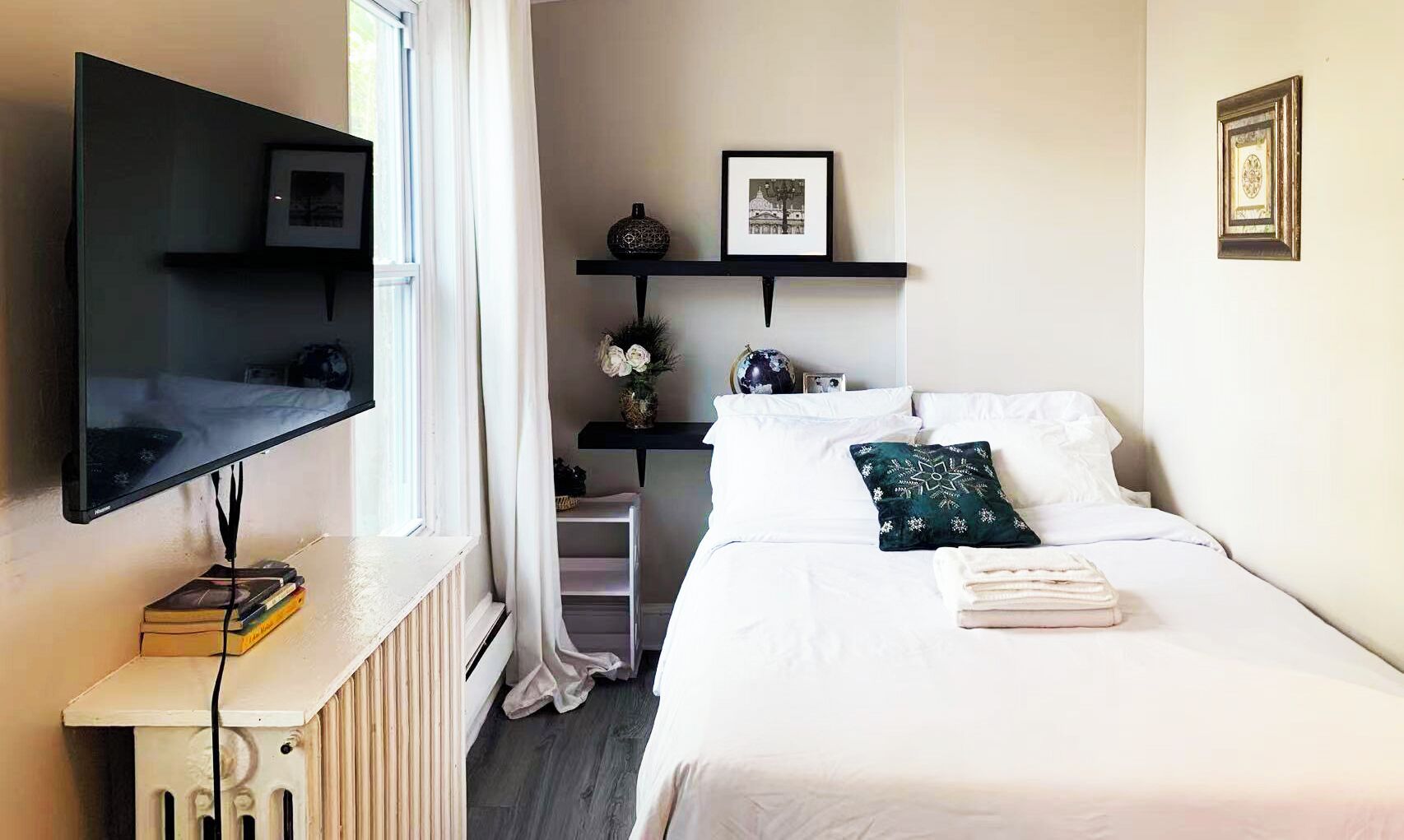 A hotel room with a bed, a TV mounted on the wall, a radiator, and a window with curtains.