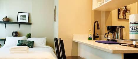 A hotel room with a bed, a small table, a chair, a microwave, a coffee maker, and a shelf with various items.