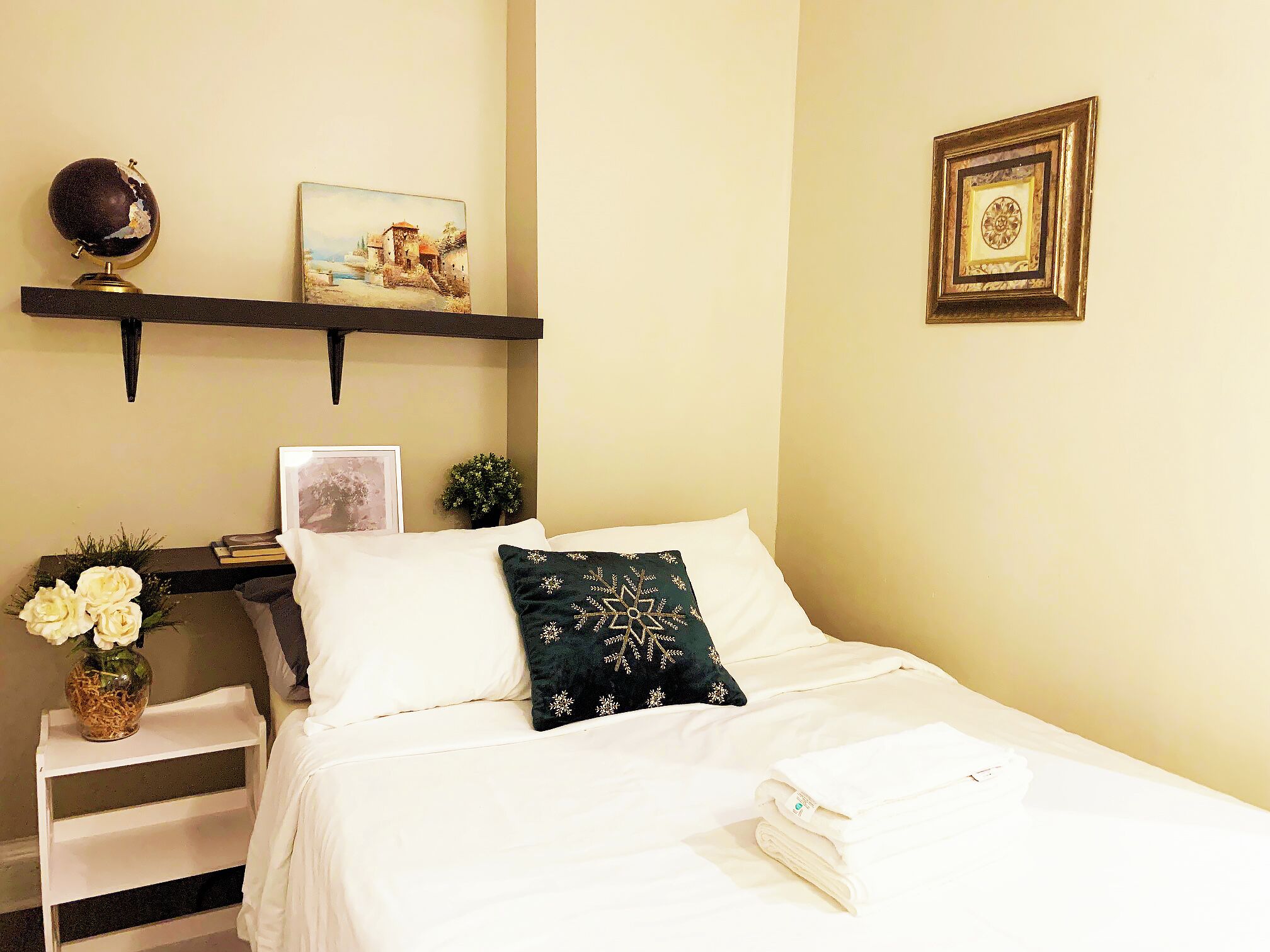 A neatly made bed with white linens and a decorative pillow, a small bedside table with a vase of flowers, a globe, and a framed picture on a shelf above the bed.