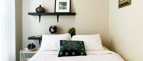 A neatly made bed with white linens, a green decorative pillow, and a stack of white towels. There are two wall-mounted shelves with decorative items and a framed picture.