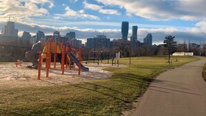 Un parque infantil con un tobogán y estructuras para escalar, un sendero pavimentado y un horizonte urbano de fondo.